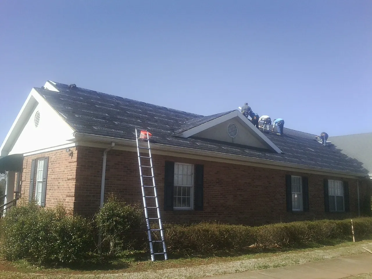 Crew tearing shingles off a brick ranch roof with a ladder set — Emergency Roof Repair work by our crew in Homeland