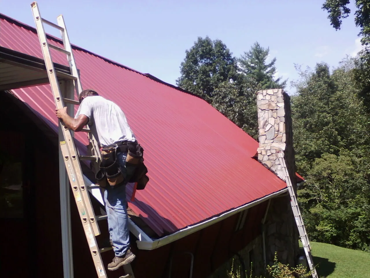 Roofer on a ladder beside a red metal roof and stone chimney — Soffit & Fascia Repair by Roofing Brothers in Homeland